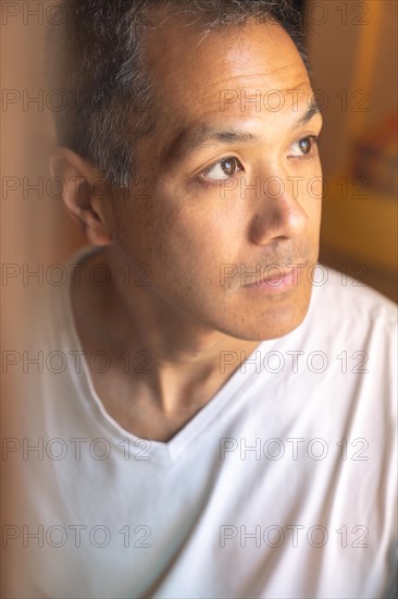 Vertical portrait of young Japanese man gazing out of a window with a pensive expression. Concept related to mental health, contemplation, introspection, or melancholy