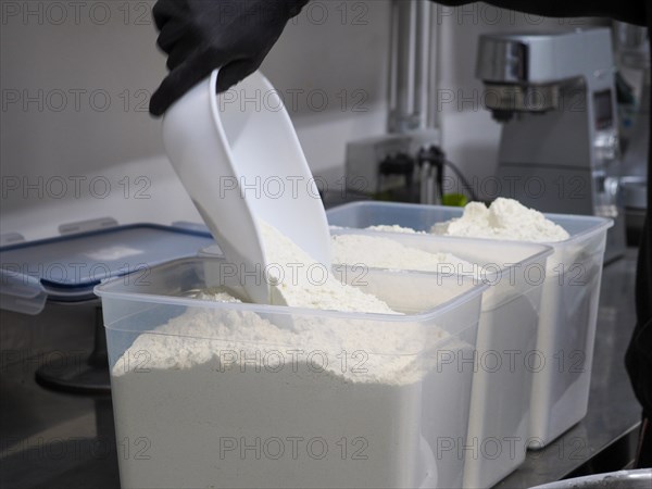 Pouring white flour from a large container with a scoop in a commercial kitchen setting