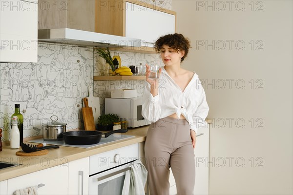 Young woman wearing a white shirt tied at the waist and beige trousers standing in the kitchen with a glass of filtered clean water