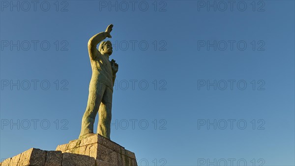 Statue of a man pointing to the sky, standing on a pedestal, bronze statue of a waving sailor in memory of the souls lost at sea, in front of a clear blue sky, Gythio, Mani, Peloponnese, Greece, Europe