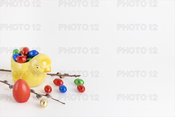 Colourful chocolate eggs in a yellow ceramic chick next to a palm kitten and candle, white background, copy room