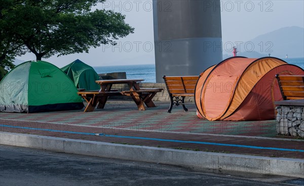 Tents set up on walkway next to picnic table and benches with the ocean and a bridge support column in the background in South Korea