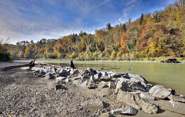 An angler sits on large stones by a river in autumn, with a castle perched on a wooded hill in the background, Salzach, Burghausen, Upper Bavaria, Bavaria, Germany, Europe
