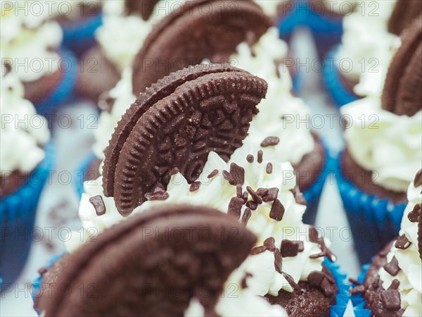 Close-up of a large group chocolate cupcakes with white cream frosting, dark chocolate cookie topping and sprinkles in blue liners