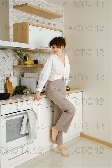 Young barefoot woman standing by the countertop of a well-lit and elegantly designed modern kitchen and looking at frying pan on electric stove