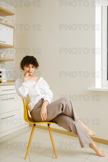 Young barefoot woman with curly hair wearing casual clothes sits in the kitchen propping chin