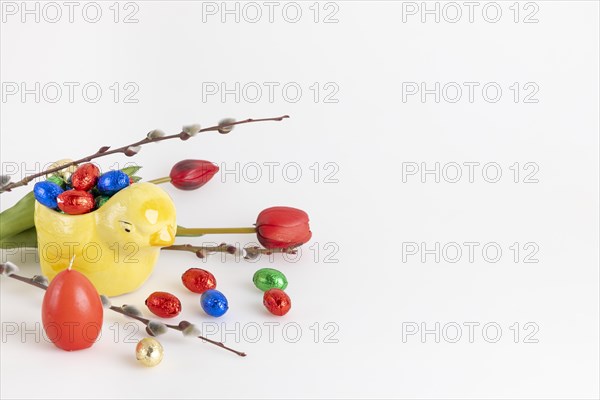 Colourful chocolate eggs in a yellow ceramic chick next to palm catkins, tulips and candle, white background, copy room