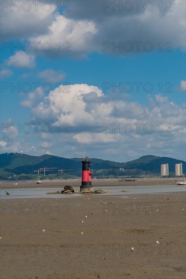 Red and black lighthouse on tidal flats during low tide with mountains and cloudy blue sky in background in Yeosu, South Korea, Asia