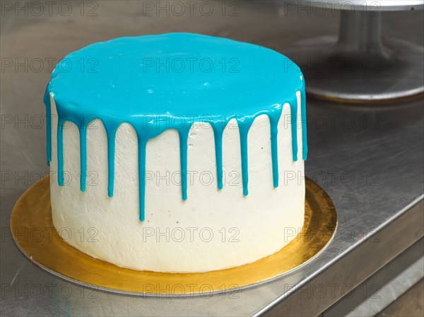 A white frosted cake with vibrant blue dripping icing on a stainless steel surface ready to be decorated and topped for a birthday celebration