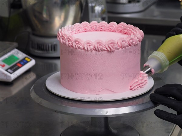 Pastry bag with green nozzle applying pink icing on a cake held by hands in black gloves