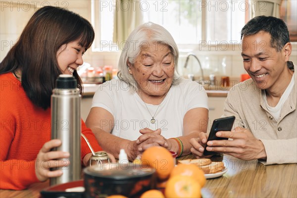 Adult children teach their elderly mother of Japanese origin how to use a cell phone while sitting in the kitchen drinking mate. Argentine-Japanese family