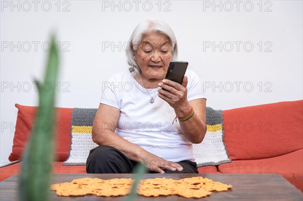 Older woman of Japanese ethnicity with grey hair looking at her mobile phone screen, sitting at home. Online communication using a smartphone