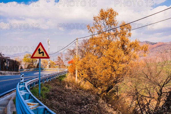 Warning sign next to guardrail on curvy mountain road with cloudy sky in background in South Korea