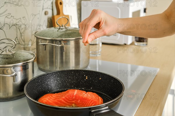 Unrecognizable woman adds salt on piece of trout steak in frying pan