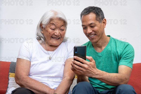 Adult son teaching his elderly mother of Japanese origin how to use a mobile phone. Elderly mother and Latin-Japanese son looking at the phone and smiling, enjoying time together at home