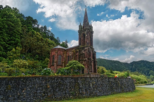 Old gothic church Capela de Nossa Senhora das Vitorias surrounded by trees and a stone wall against a dramatic cloudy sky, Furnas Lake, Furnas, Sao Miguel, Azores, Portugal, Europe