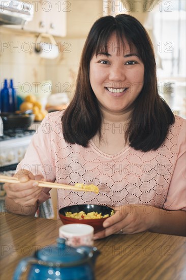 Vertical portrait of Asian woman sitting at home eating rice with chopsticks looks and smiles at camera