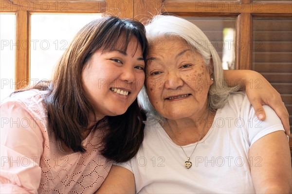 Close up of Japanese adult daughter hugging her grey haired mother smiling and looking at the camera