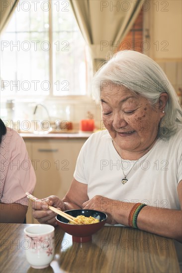 Portrait of an elderly Japanese woman with gray hair eating rice with chopsticks. Vertical portrait bright background
