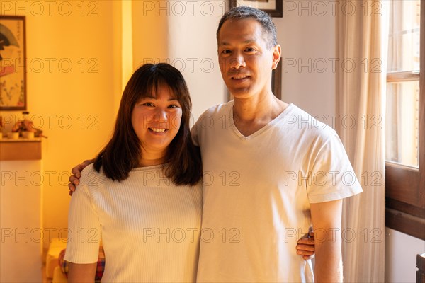 Adult siblings standing hugging each other of Japanese ethnicity, smiling and looking at the camera. Horizontal portrait indoors with warm light
