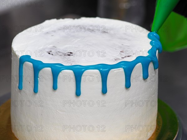 A white frosted cake in the process of being decorated with blue icing with a filling bag with large nozzle