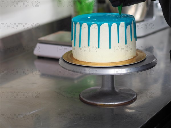 A frosted cake with white frosting being decorated with blue icing on a kitchen counter by pastry chef wearing black latex gloves