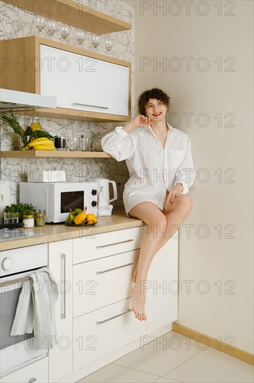 Barefoot joyful young woman wearing a white shirt sitting on the countertop of a modern kitchen