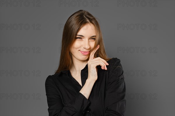 Playful young woman winking with tricky smile on grey studio background