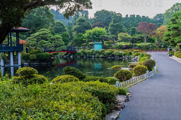Paved walkway next to koi pond with a footbridge leading to gazebo in manicured public park in South Korea