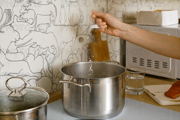 Unrecognizable woman use spoon to add salt to a pot with boiling water on the electric stove