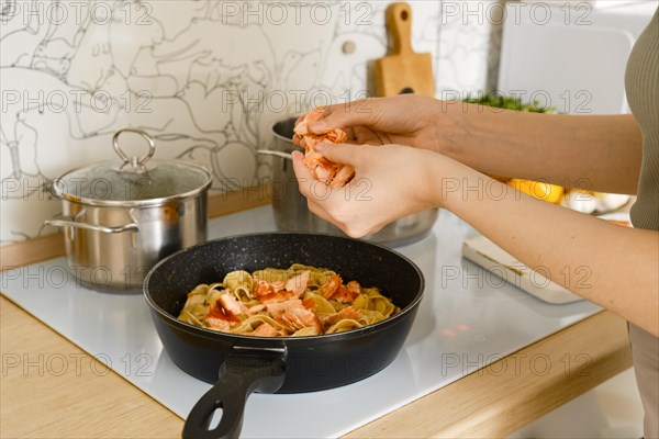 Hands of woman breaking fried salmon into pieces and crumbles it into a frying pan with pasta