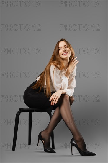 Smiling woman in pencil skirt and blouse leaning forward while sitting on a chair on grey studio background