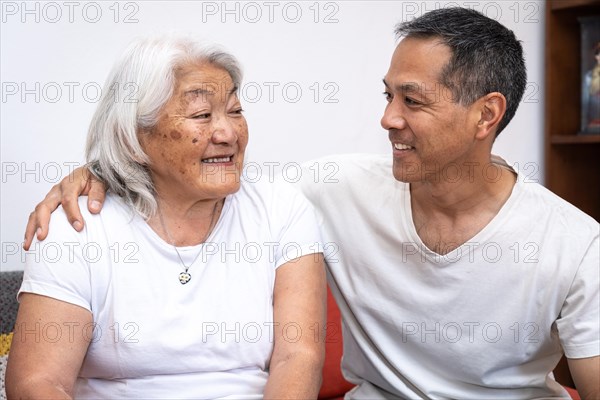 Asian mother looking at her adult son sitting on a sofa at home, happy family lifestyle concept