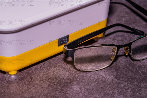 Closeup of pair of wire framed glasses in front of ultrasonic cleaner machine on black textured background