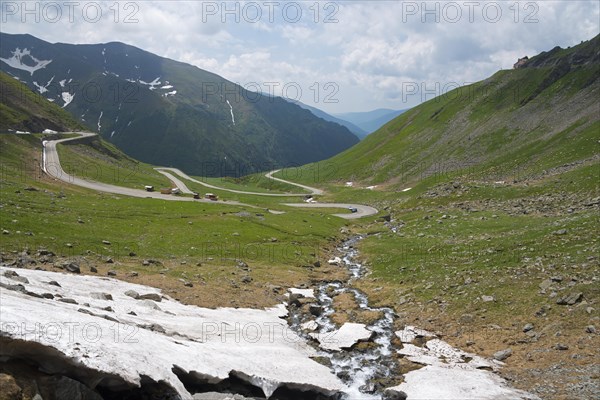 A winding road meanders through a green valley with snowfields, Capra River, mountain road, Transfogarasan High Road, Transfagarasan, TransfagaraÈ™an, FagaraÈ™ Mountains, Fagaras, Transylvania, Transylvania, Transylvania, Ardeal, Transylvania, Carpathians, Romania, Europe