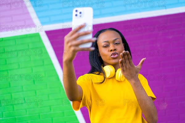 African woman blowing a kiss while taking a selfie against a colorful urban wall outdoors