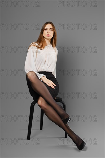 Stylish young woman with long legs in black tights sits on a chair on grey background