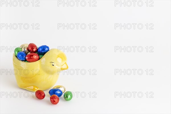 A yellow ceramic chick filled with a selection of colourful chocolate eggs, white background, copy space