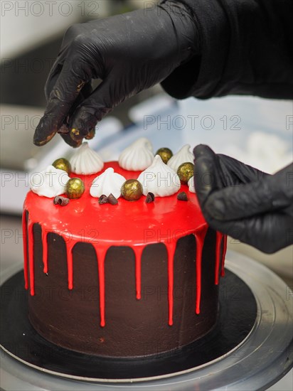 A black gloved hand adding a gold-colored ball to garnish a red-glossy drip cake