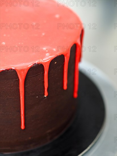 A dark chocolate frosted cake with glossy red dripping icing on a stand