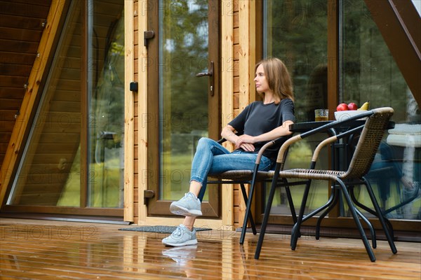 Cute woman enjoys fresh air after the rain sitting on terrace of her forest cabin