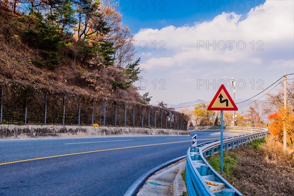 Warning sign next to guardrail on curvy mountain road with cloudy sky in background in South Korea