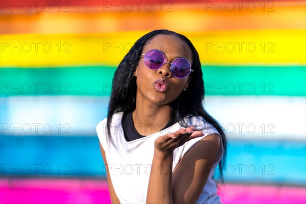 African woman with sunglasses gesturing blowing a kiss in a multicolored vivid urban background