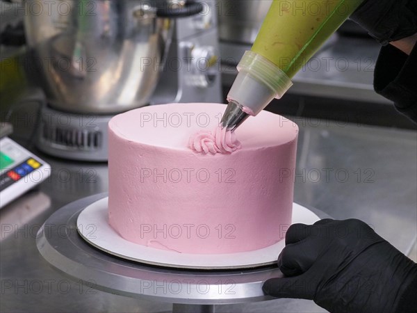 Close-up of a confectioner's piping bag adding pink frosting to decorate a cake with swirls, wearing black gloves