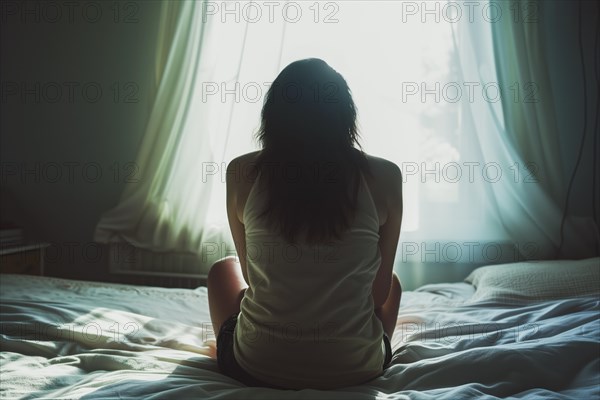 A young woman sits huddled on a bed in the morning sunlight, symbolising depression and abuse, AI generated, AI generated