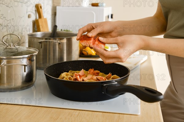 A woman breaks fried salmon into pieces and crumbles it into a frying pan with pasta