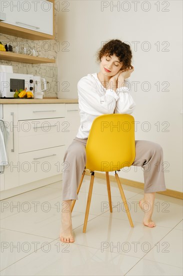 Dreamy young woman sits on chair in the kitchen with closed eyes
