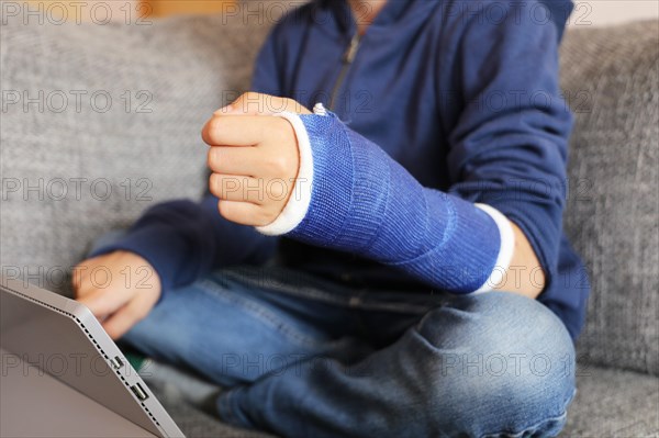 Boy with arm in plaster sits on the sofa with a tablet