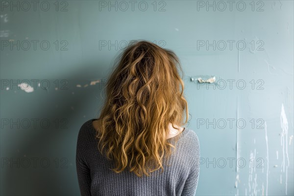 A young woman stands in front of a green wall and you see her from behind, her wavy hair in focus, symbolising depression and abuse, AI generated, AI generated