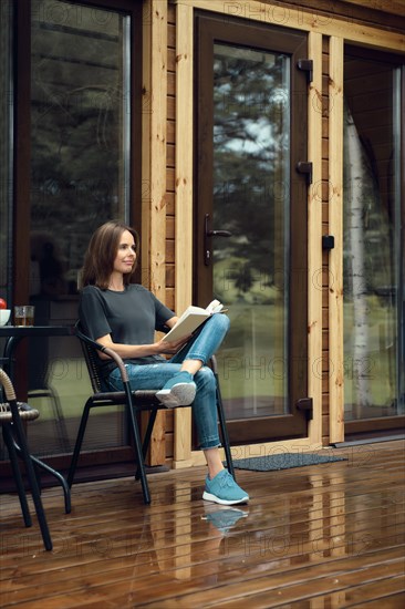 Cute woman in casual clothing sits reading on a wet terrace of a log cabin after the rain, enjoying fresh air of woods